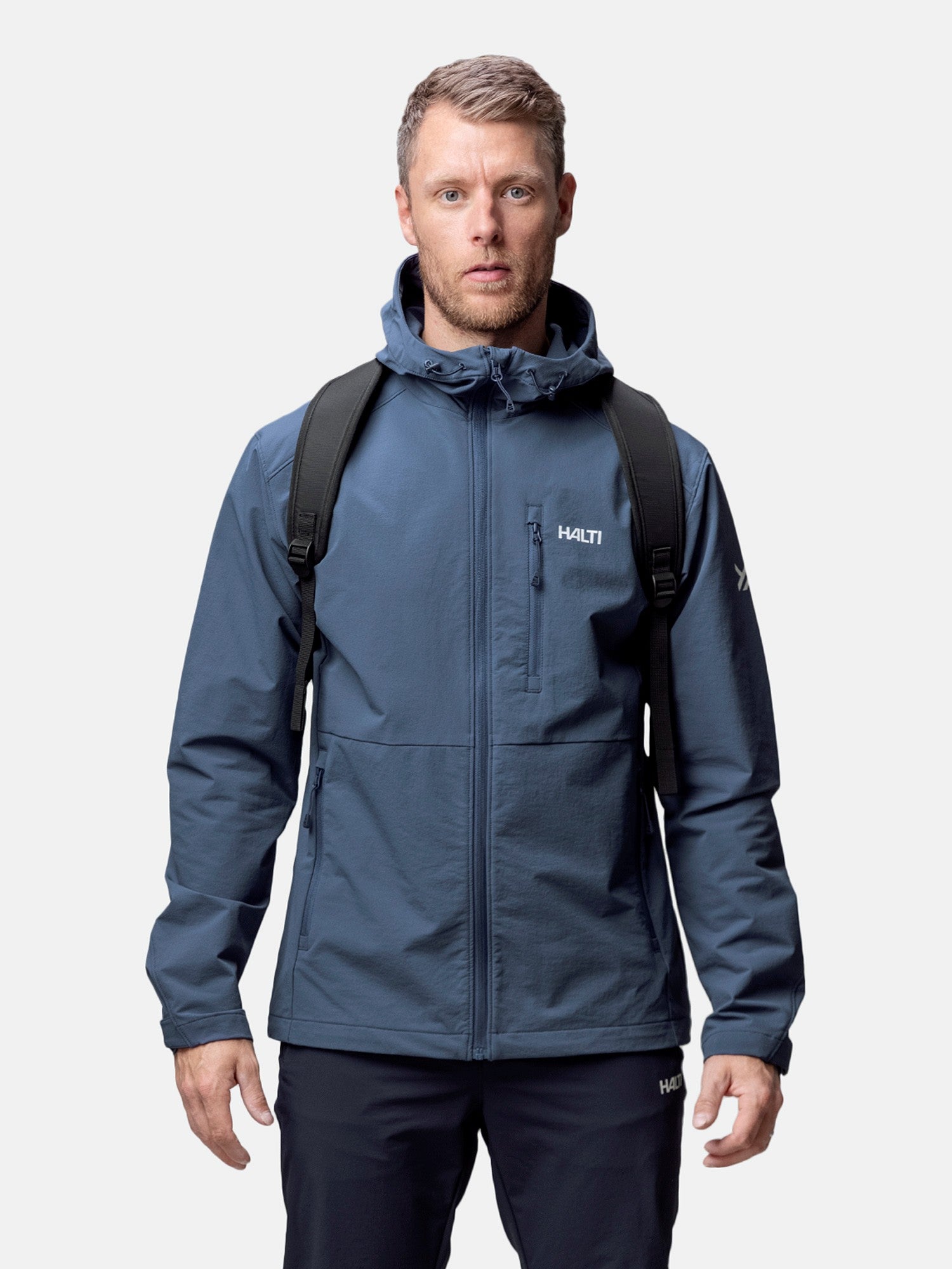 En man i Halti Finholma X-stretch Jacka Herr och svart ryggsäck står vänd mot kameran, redo för friluftsliv.