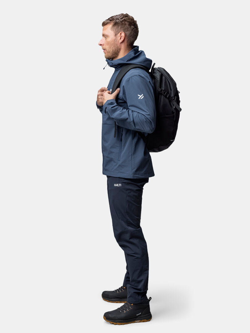 Man i Halti Finholma X-stretch Jacka Herr, byxor, svarta sneakers och ryggsäck står i profil mot en ljus bakgrund.