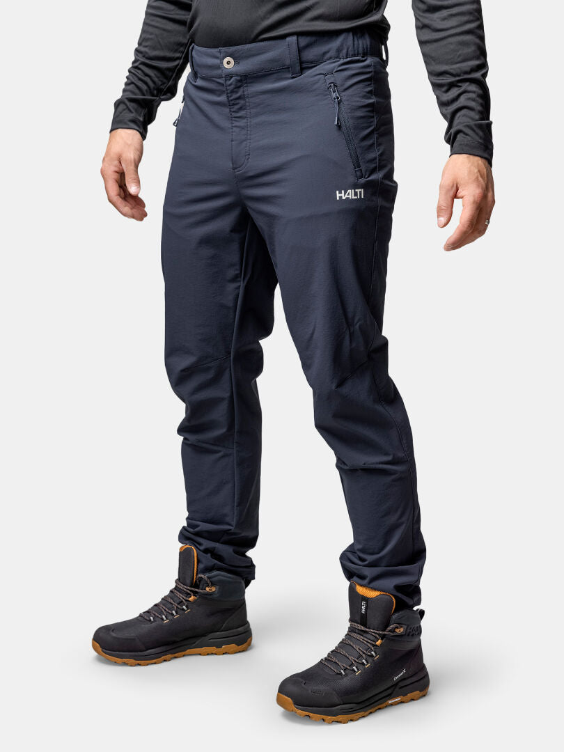 En person i Halti Finholma X-stretch Byxor Herr, svarta stövlar och en långärmad skjorta står mot en enfärgad bakgrund.