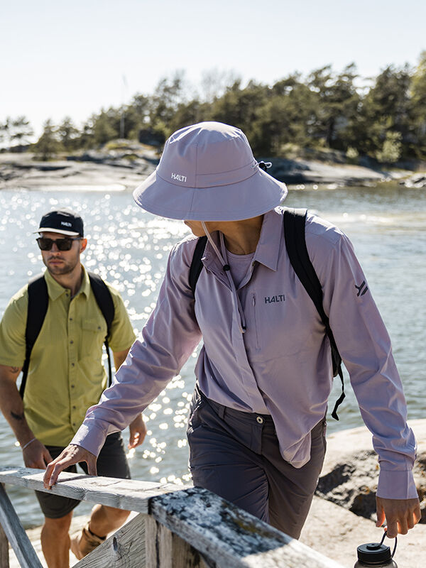 Två personer i Halti Samoy Långärmad Funktionsskjorta Dam och ryggsäckar går vid en sjö, träd och klippor bakom dem.
