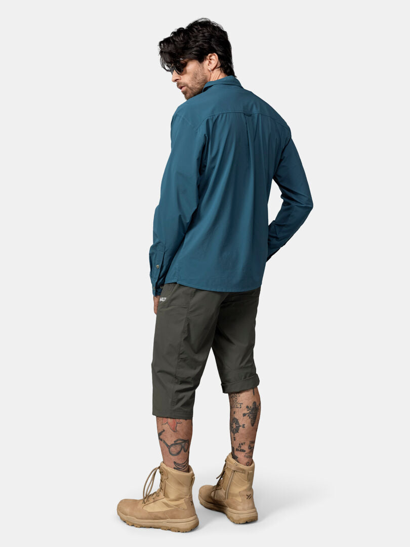 En tatuerad man vänder sig bort i en blågrön Halti Samoy Långärmad Funktionsskjorta Herr, mörka shorts och solbruna stövlar.