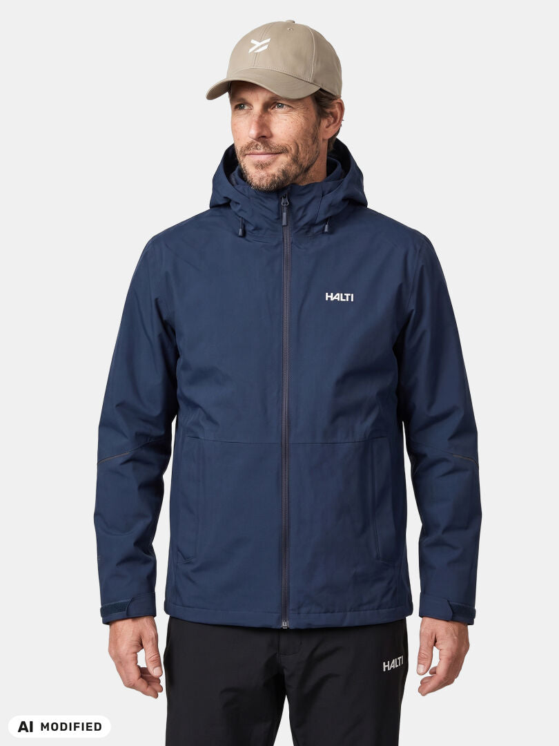 En man bär en Halti Loito 3in1 DX Jacka Herr i blått och en beige keps. Han står framför en helt vit bakgrund och blickar rakt fram.