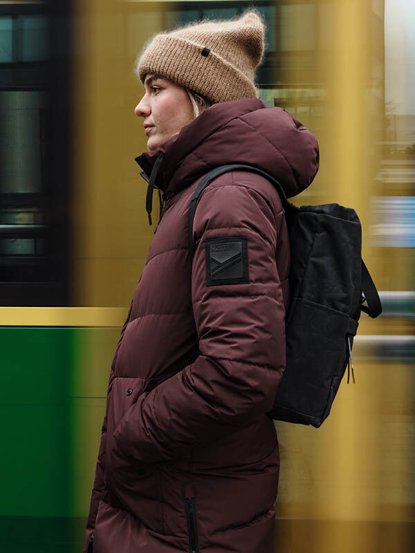 En person i brun Halti Linjat Parka Jacka Dam och brun mössa står med en svart ryggsäck när en spårvagn passerar.