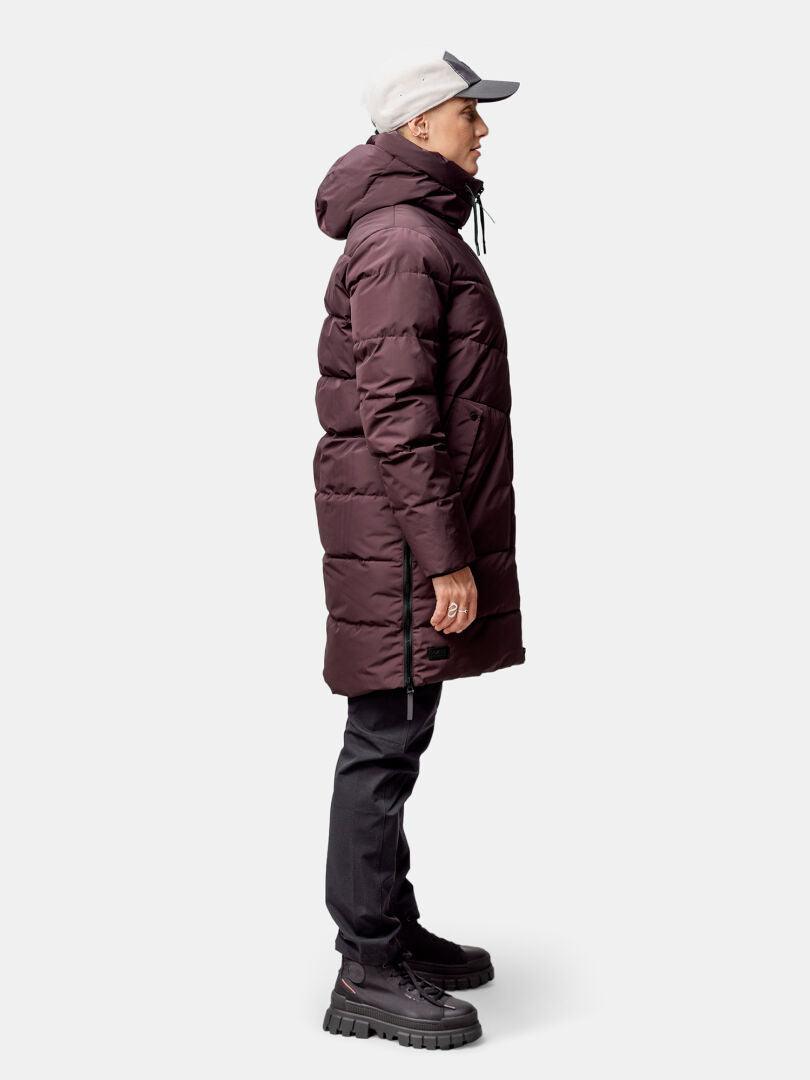 En person i en rödbrun Halti Linjat Parka Jacka Dam står i profil mot en enfärgad bakgrund.