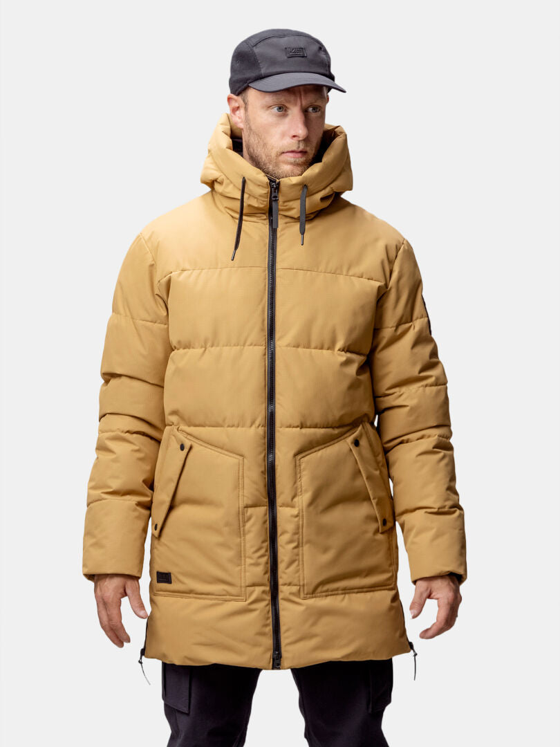 En man i brun Halti Linjat Parka Jacka Herr, svart keps och svarta byxor står mot en ljus bakgrund.