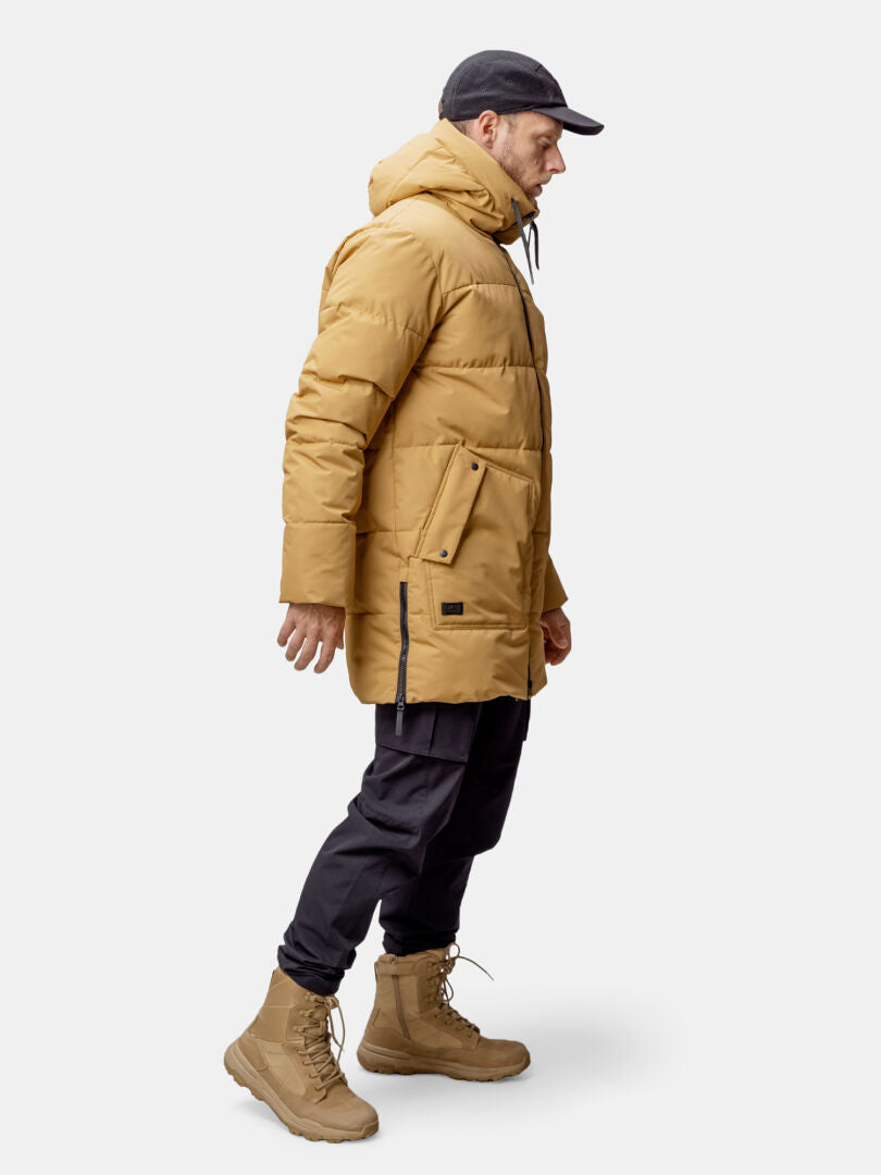 En man i Halti Linjat Parka Jacka Herr, svarta byxor och beigefärgade stövlar står mot en ljus bakgrund.