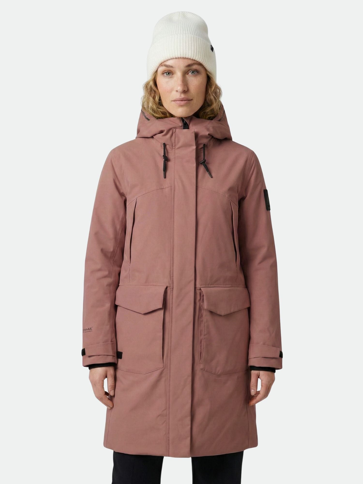 En kvinna i en Halti Bergga DX Vinterjacka Dam och en vit stickad mössa står mot en enfärgad bakgrund.