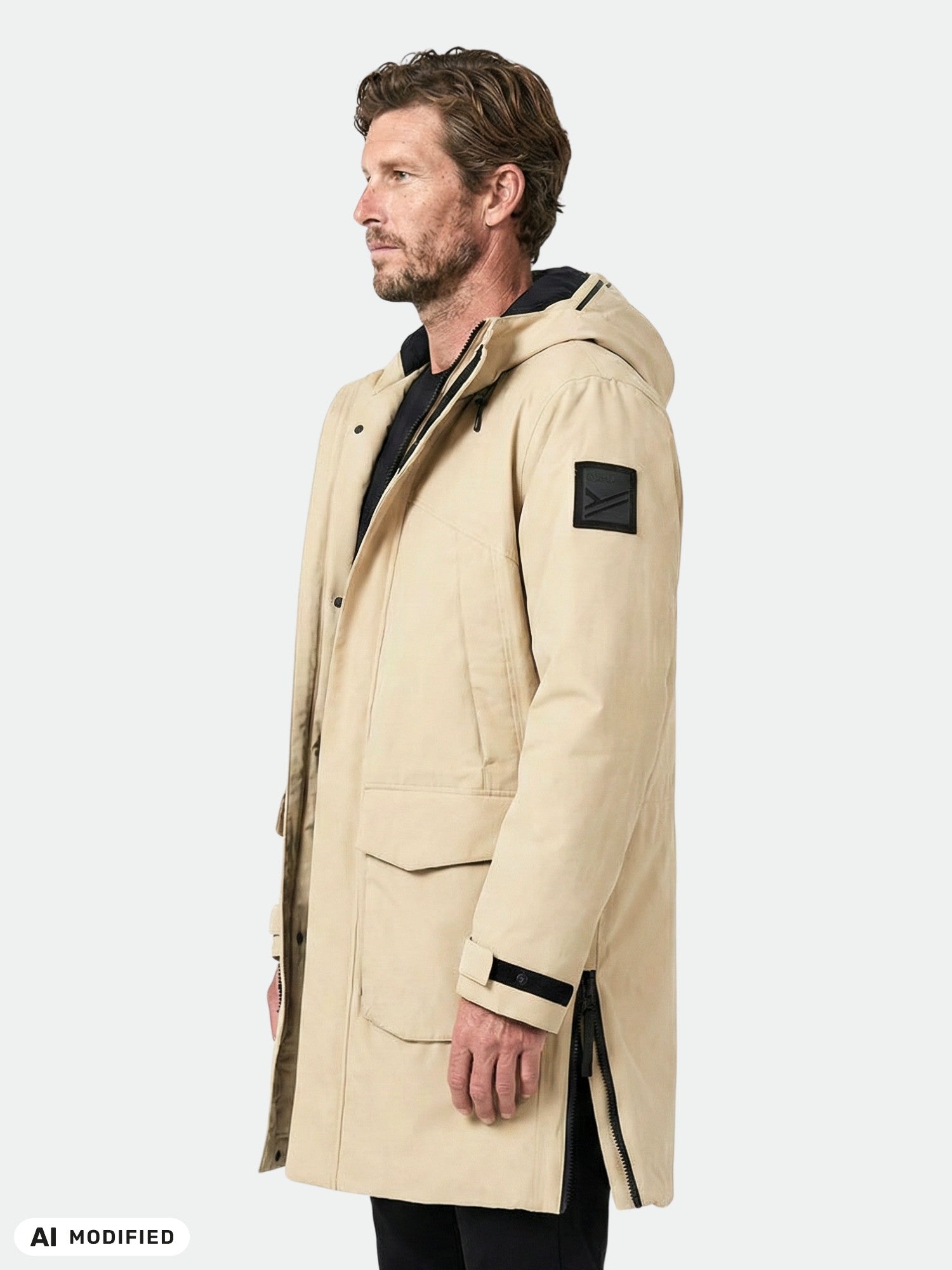 En man i en beige Halti Bergga DX Vinterjacka Herr med svart lapp på ärmen, stående i profil mot en ljus bakgrund.