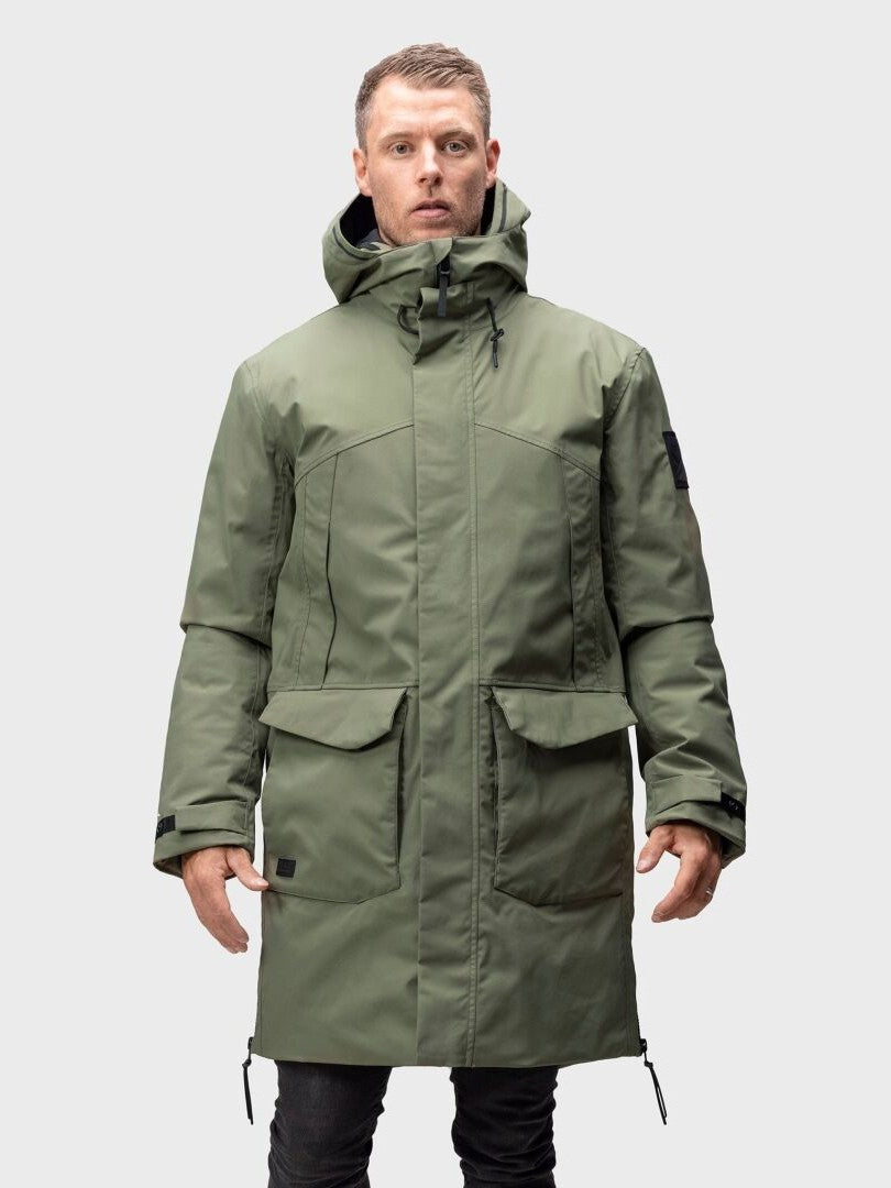 En person bär en grön Halti Bergga Herr DrymaxX Vinterjacka med stora fickor, stående mot en enfärgad bakgrund.