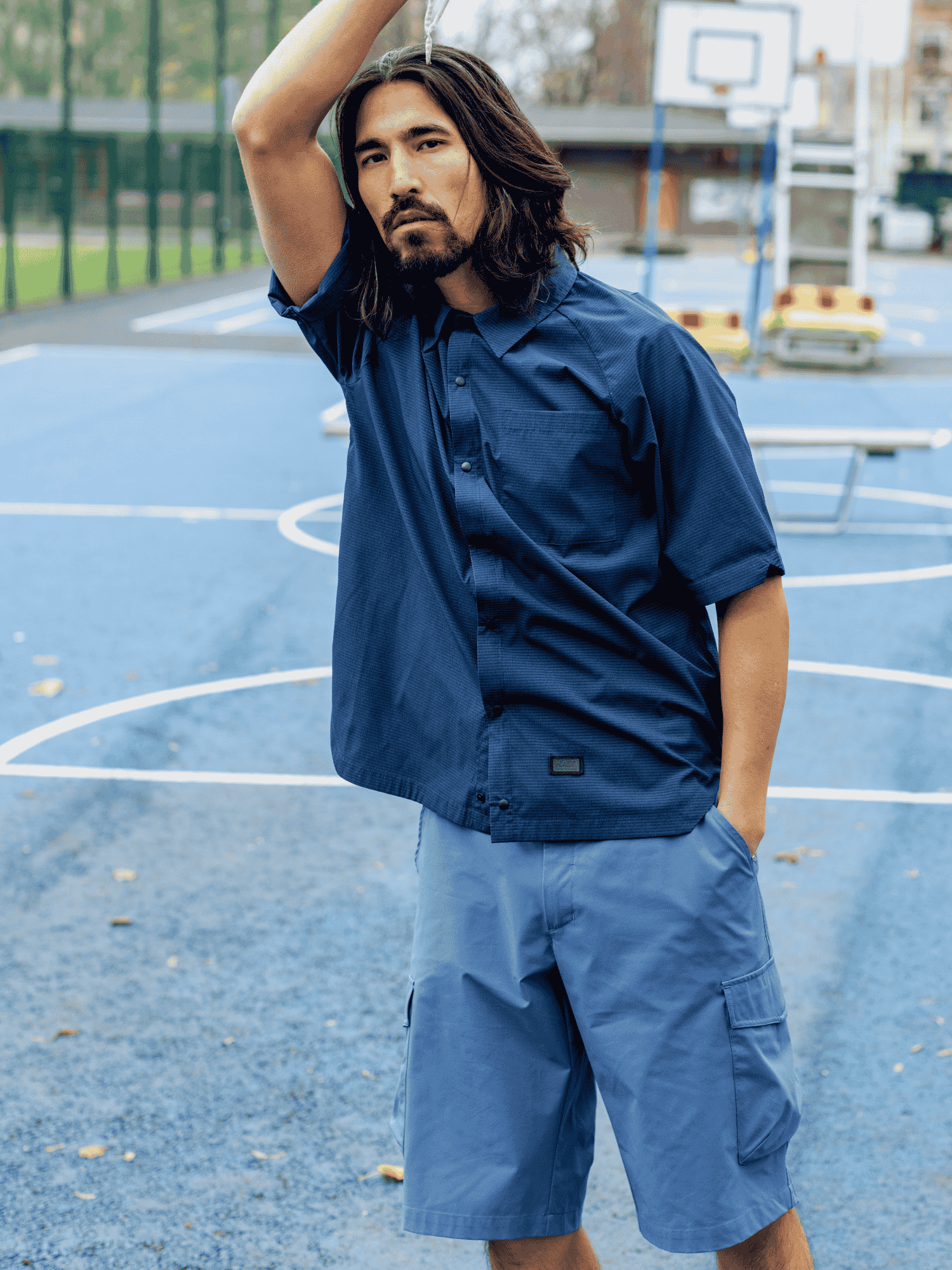 Man i en mörkblå Halti Baana Unisex Skjorta och ljusblå cargoshorts står på en basketplan utomhus.
