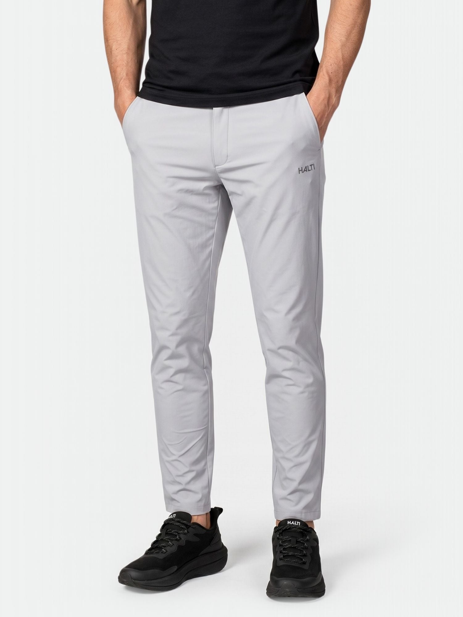 En person bär Halti Drive X-stretch Chinos i ljusgrått, en svart skjorta och svarta sneakers, händerna i fickorna.