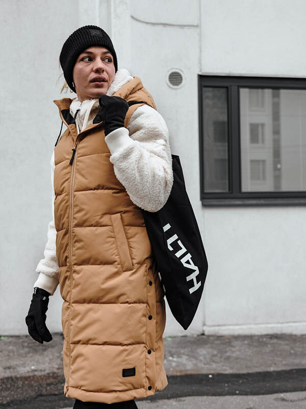 Person i Halti Penger Puffer Väst Dam och svart mössa står utomhus och håller i en svart Halti-väska.