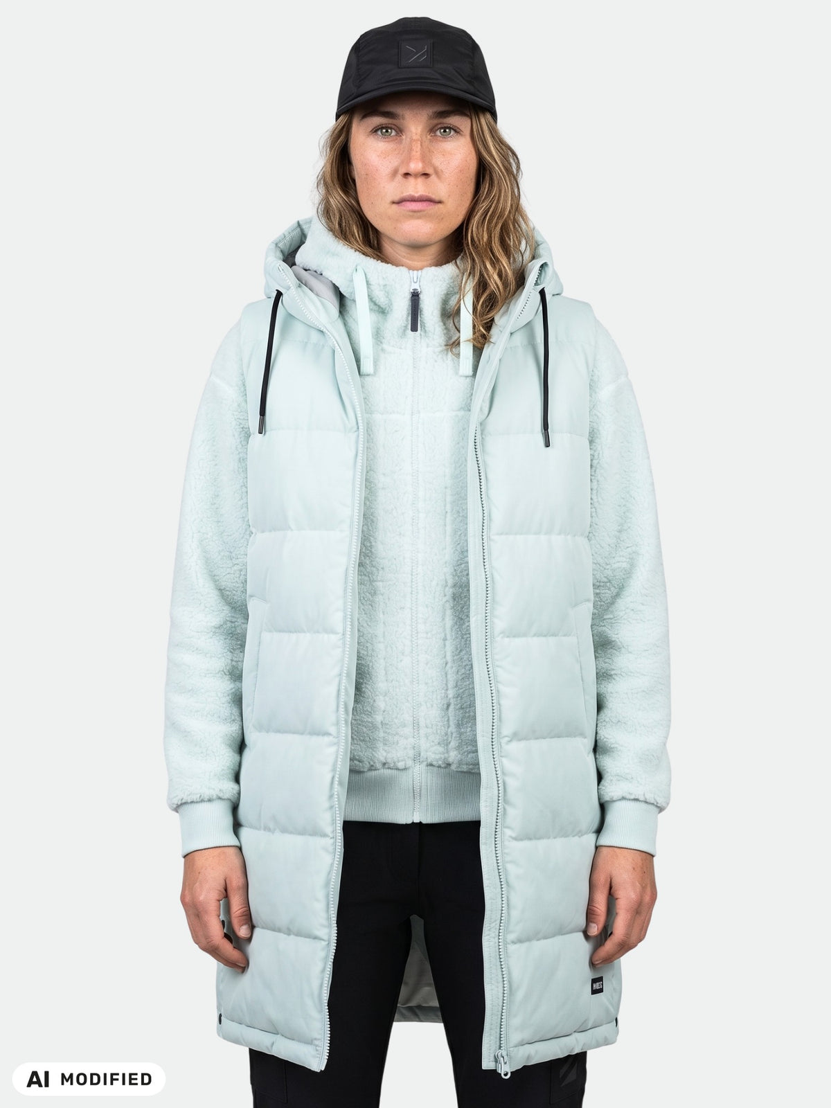 En person klädd i en Halti Penger Puffer Väst Dam, en huvtröja och en svart keps står framför en enfärgad bakgrund.