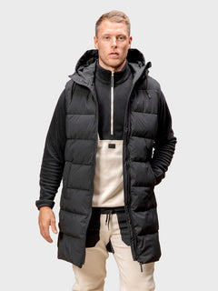En man bär en Halti Penger Puffer Väst Herr över en svart-beige zip-tröja och beige byxor mot neutral bakgrund.