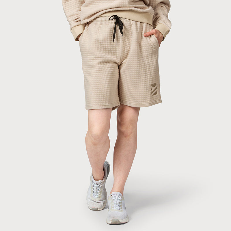 Person som bär Halti Gliffa Shorts Unisex i beige återvunnen polyester, en matchande sweatshirt och vita sneakers.