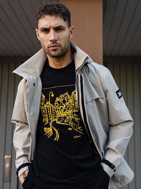 En man bär en Halti Kallio ljusgrå jacka över en Skaltsi unisex t-shirt med en gul urban grafik.