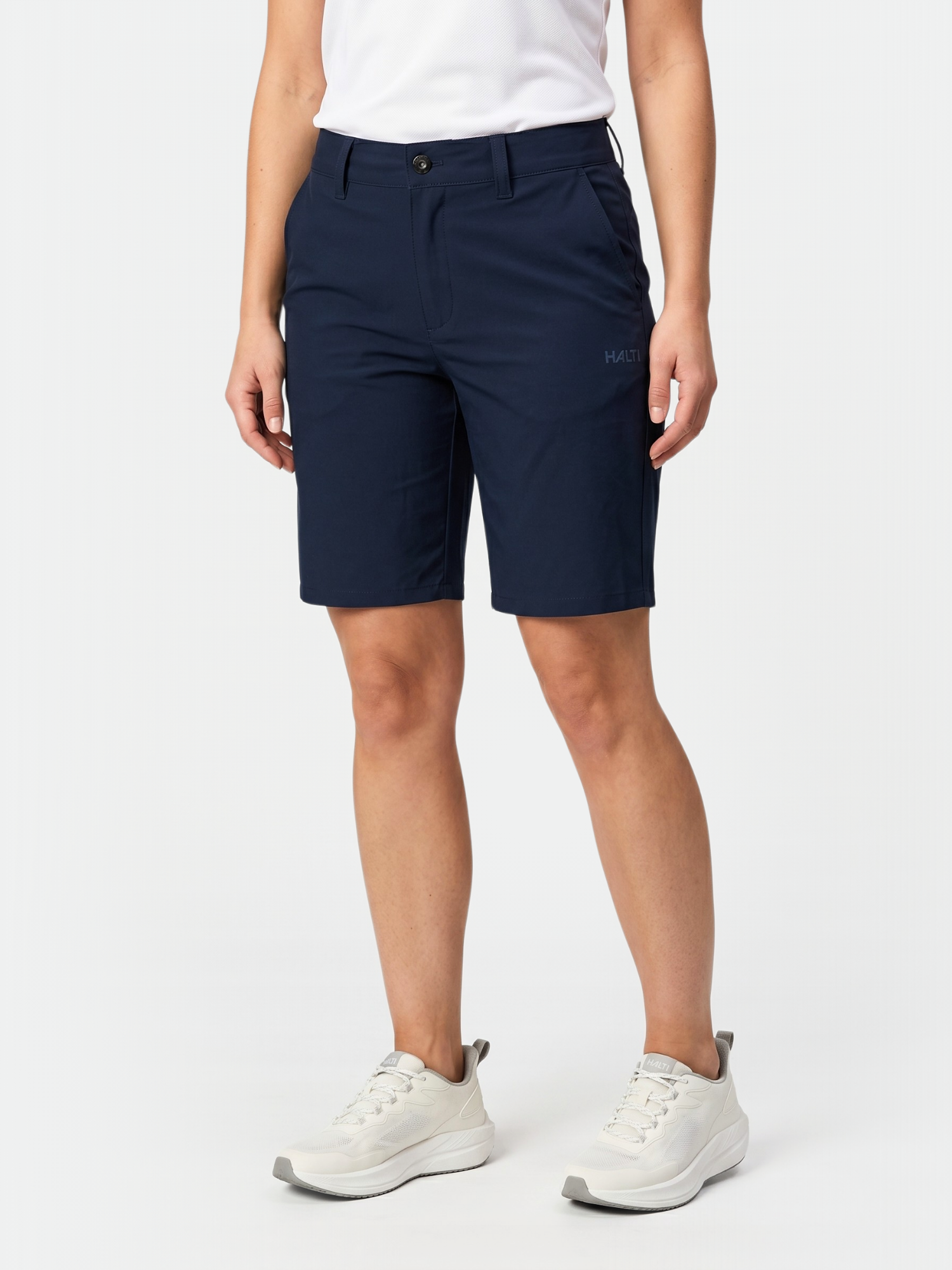 Person klädd i Halti Drive X-stretch Shorts Dam i marinblå, vit skjorta och vita sneakers mot en enfärgad bakgrund.