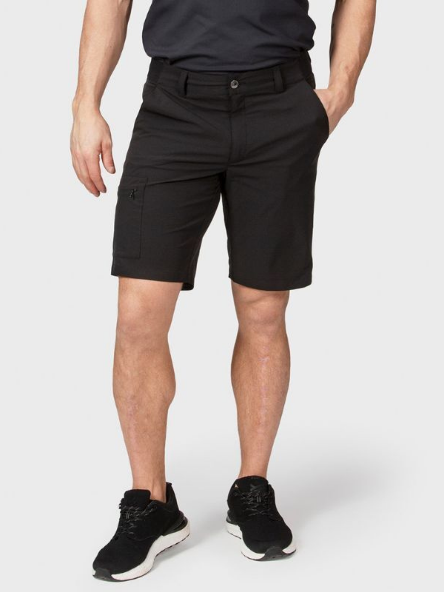 En person i Halti Drive X-stretch Shorts Herr och svarta sportskor står med händerna i fickorna mot en enfärgad bakgrund.