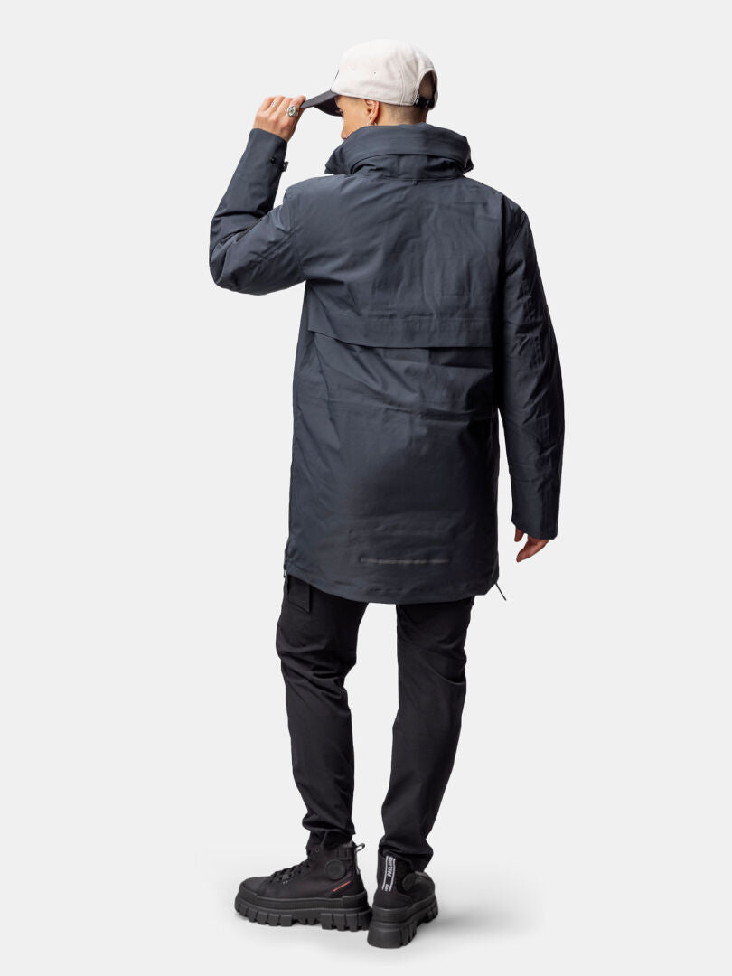 En person i Halti Synkas 3in1 DrymaxX Parka står vänd bort, klädd i svarta byxor och med en vit mössa i handen.
