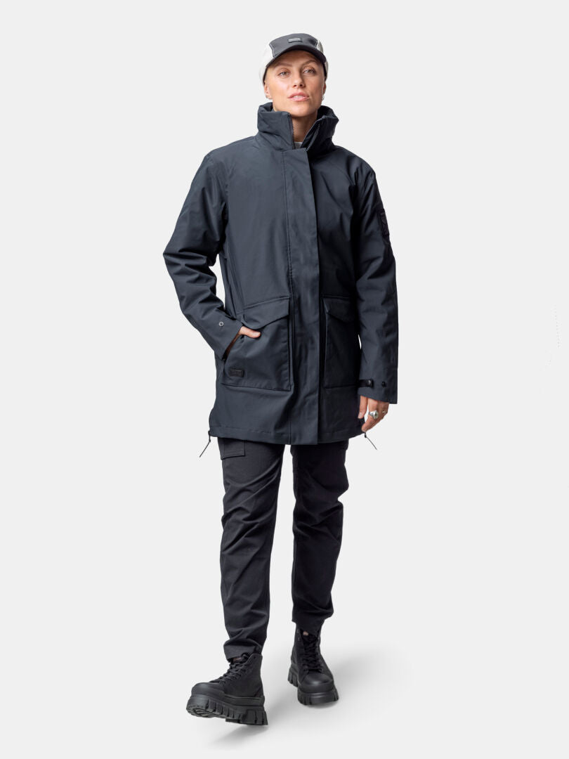 Person klädd i Halti Synkas 3in1 DrymaxX Parka Dam, svarta byxor, svarta stövlar, grå keps och med ena handen i fickan.