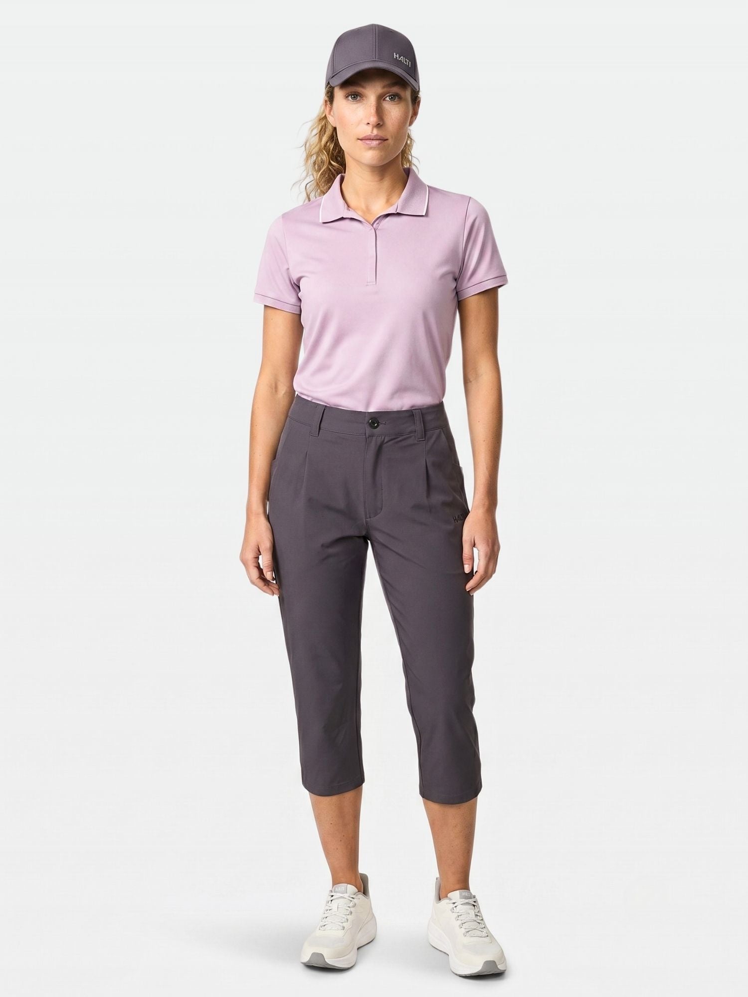 En kvinna vänder sig framåt i lila keps, UPF 50+ ljuslila polo, Halti Drive X-stretch Capribyxor Dam och vita sneakers.