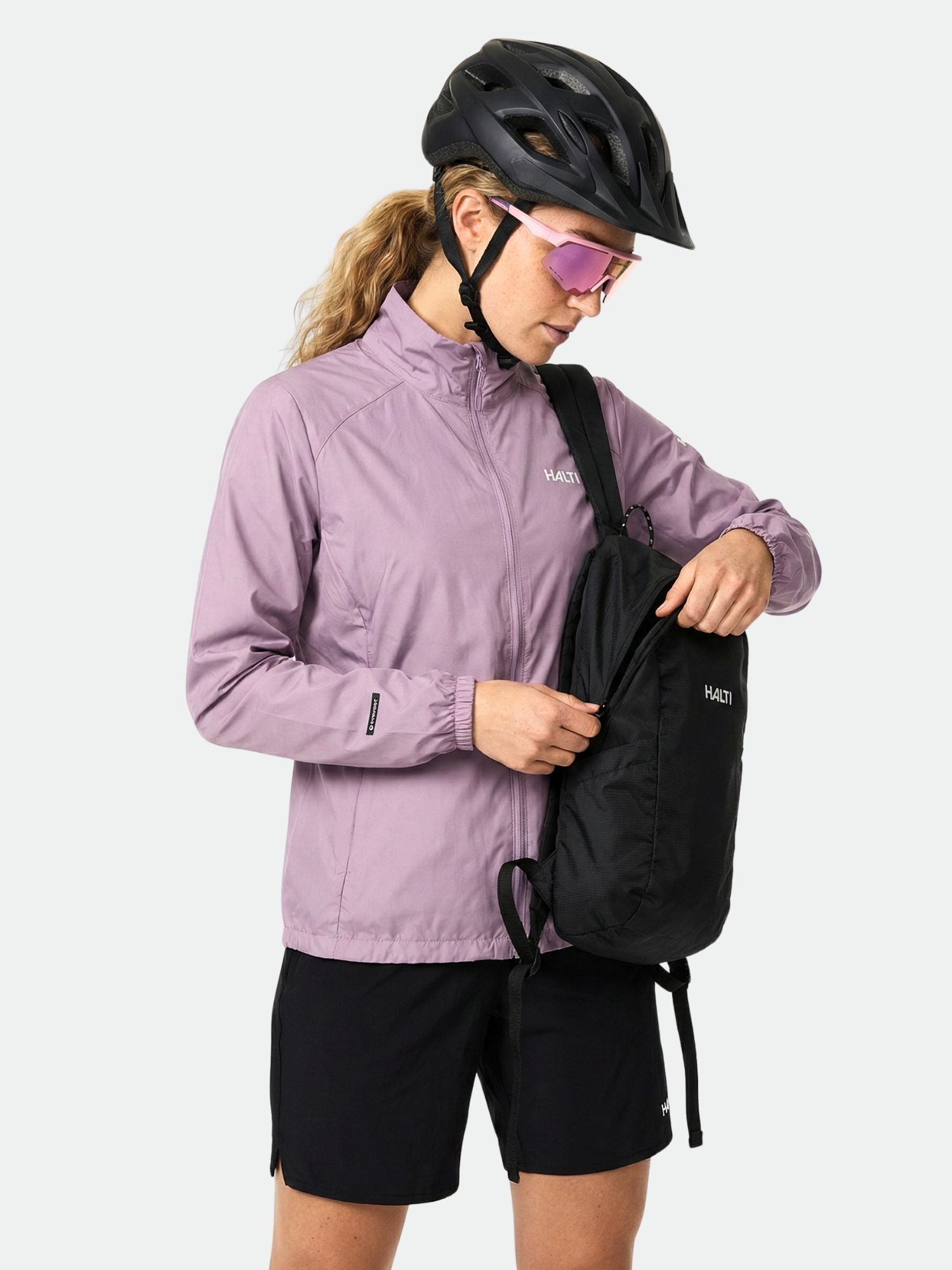 En kvinna i Halti Saime Vindjacka Dam och svarta shorts öppnar en svart ryggsäck, bär hjälm och solglasögon.