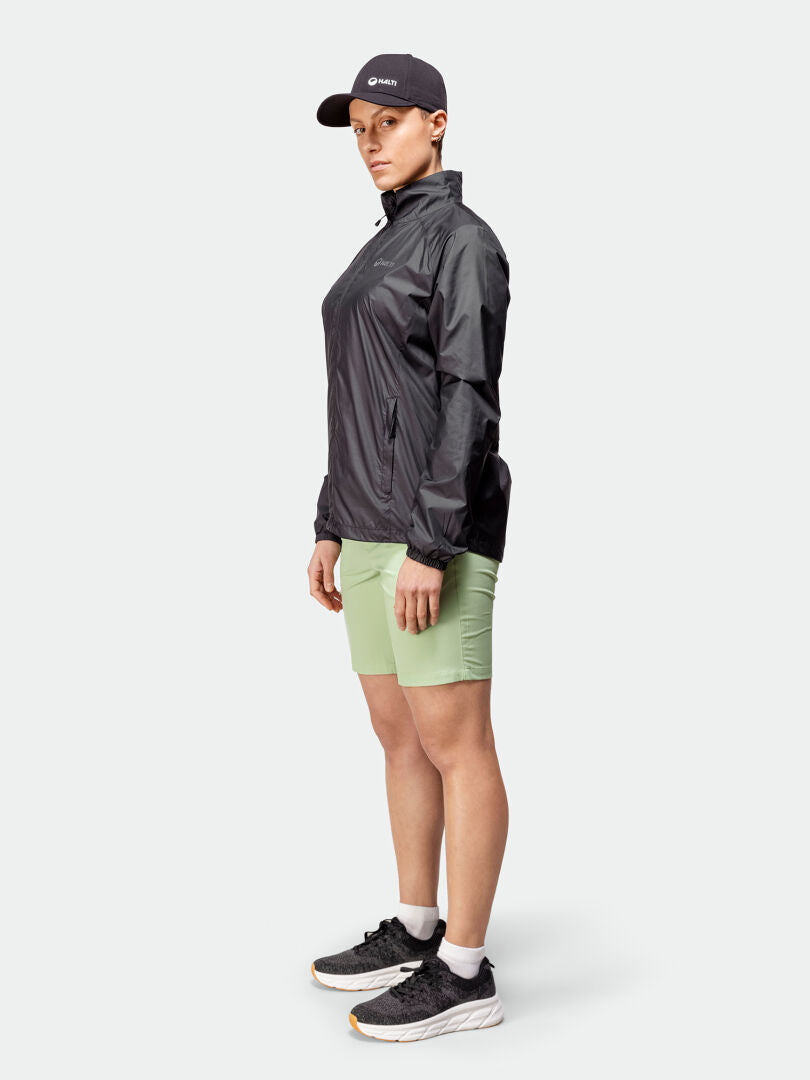 Kvinna som står snett i en Halti Saime Vindjacka Dam, gröna shorts och svarta sneakers mot en enfärgad bakgrund.