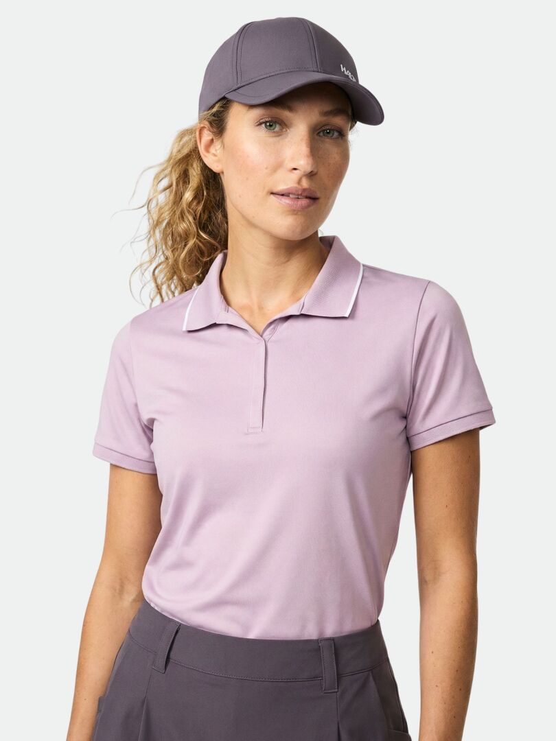 Kvinna i Halti Swingi Teknisk Pikétröja Dam av återvunnen polyesterjersey, mörk keps och byxor, mot ljus bakgrund.