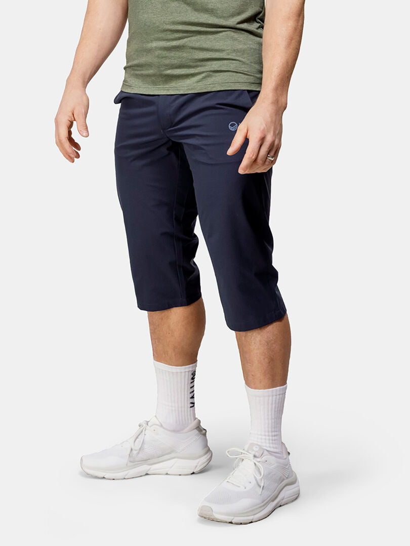 En person i en grön skjorta, Halti Drive X-stretch Capribyxor Herr, vita strumpor och sneakers står mot en enfärgad bakgrund.