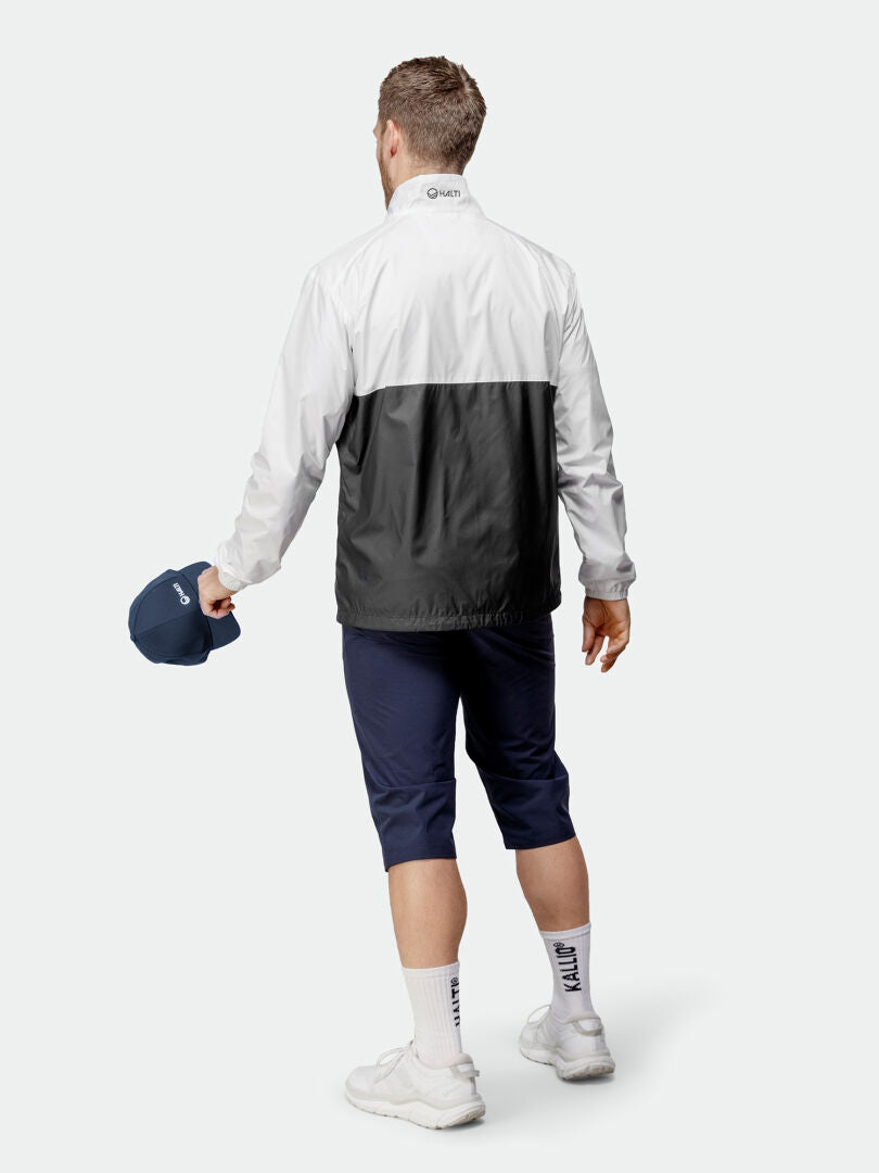 En man bär Halti Saime Vindjacka Herr, marinblå byxor, vita strumpor och sneakers och står med ryggen vänd.
