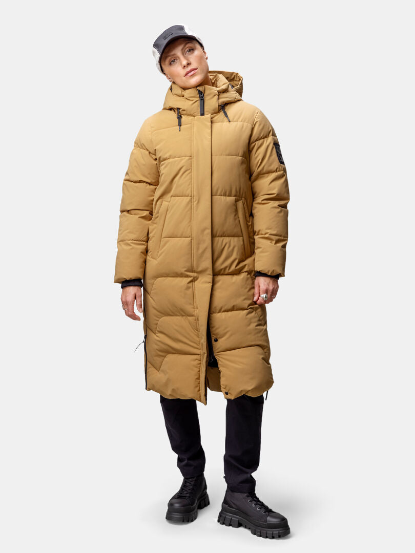 En kvinna i en Halti Brahe Puffer Long vinterjacka, svarta byxor, klumpiga skor och en ljus mössa står framför en enfärgad bakgrund.