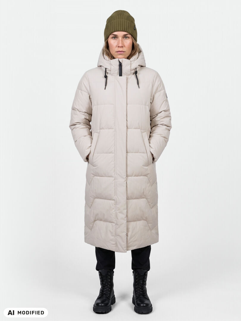 En person klädd i en lång beige Halti Brahe Puffer-vinterjacka, en olivgrön mössa, svarta byxor och stövlar framför en enfärgad bakgrund.