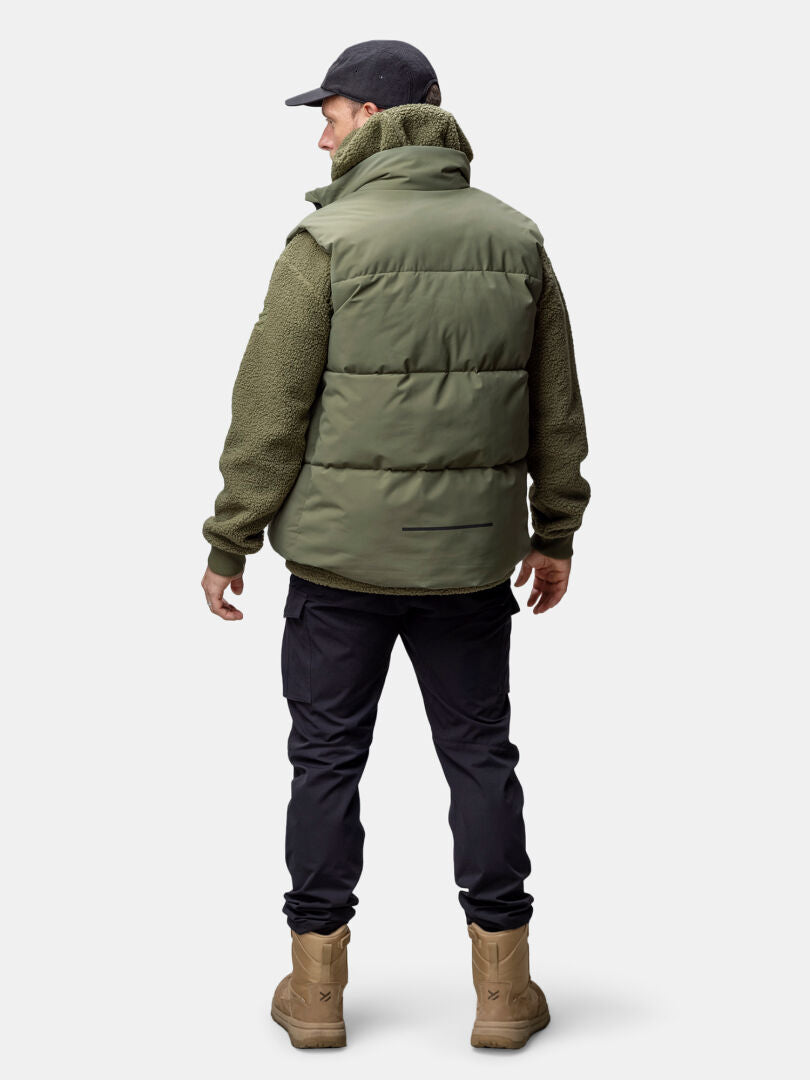 En man klädd i Halti Penger Classic Puffer Väst Herr, mörka byxor och solbruna stövlar står vänd bort från kameran.