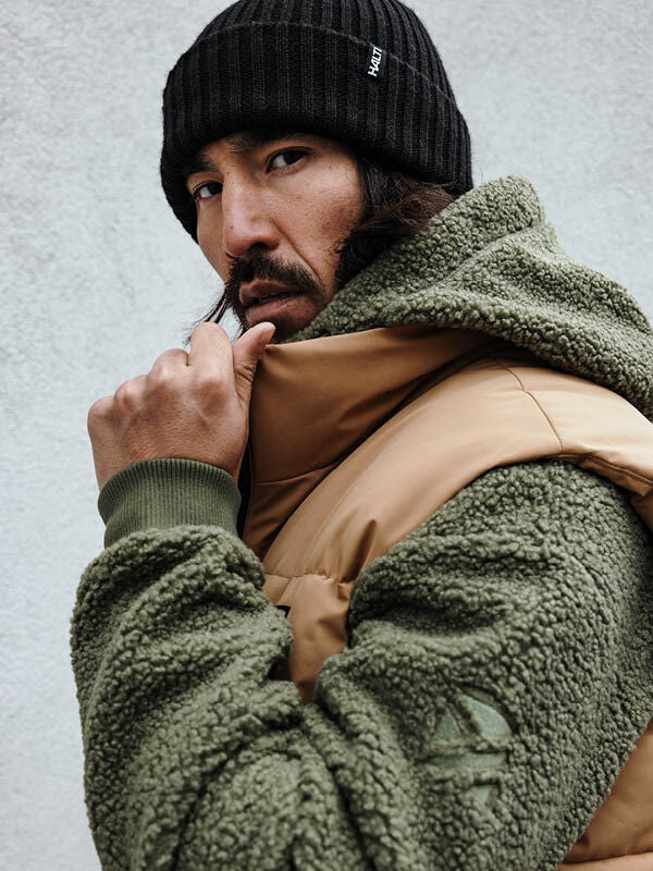 En man i en grön sherpa-jacka och en Halti Runko-mössa tittar över axeln medan han håller i kragen, mot en enfärgad bakgrund.