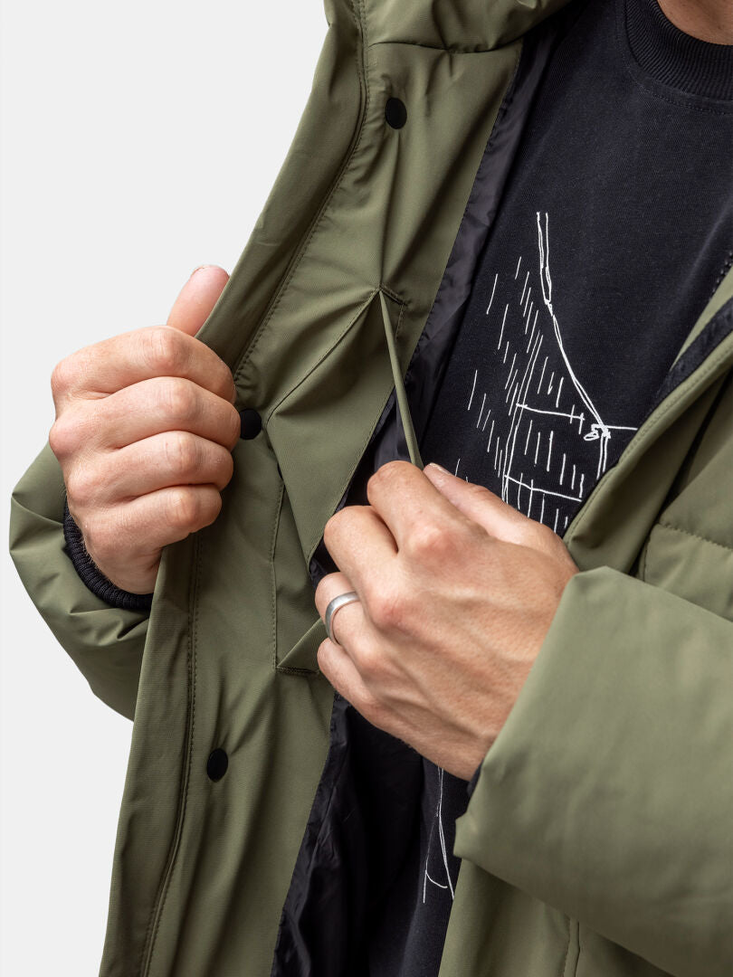 Någon justerar dragsnöret inuti en Halti Brahe Puffer Vinterjacka Herr i olivgrönt över en svart grafisk t-shirt.