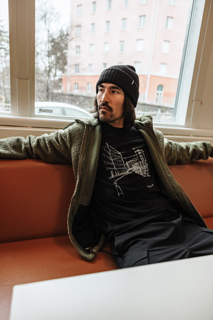 En man i grön Halti Lykky Fleecejacka Herr sitter på en orange bänk vid ett fönster och har på sig en grafisk t-shirt.
