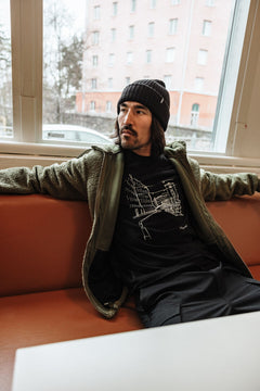 En man i grön Halti Lykky Fleecejacka Herr sitter på en orange bänk vid ett fönster och har på sig en grafisk t-shirt.