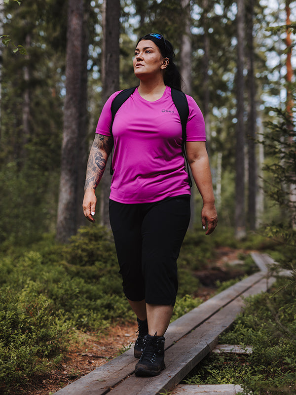 En person i rosa skjorta och Halti Drive X-stretch Capribyxor Dam Plus går på en skogsstig med en ryggsäck.