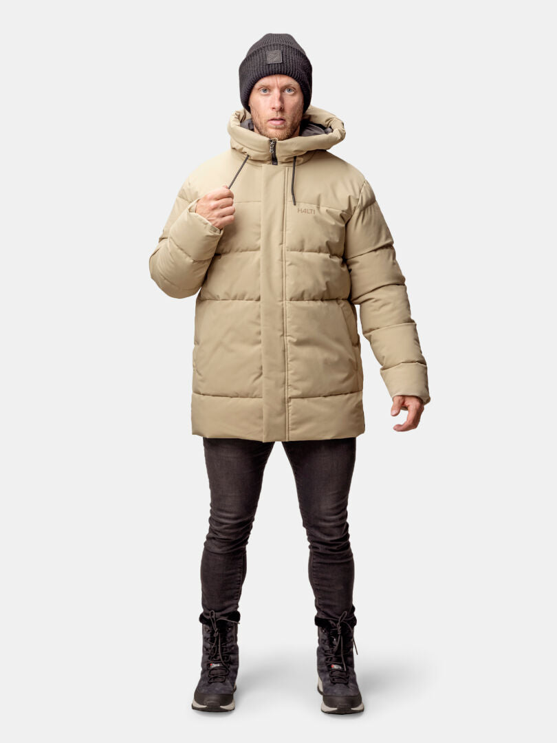 En man bär Halti Sammal Vadderad Parkajacka Herr i beige, svarta byxor, stövlar och en grå mössa.