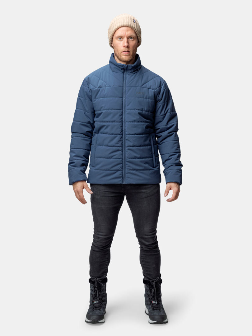 En person i Halti Haapa Vadderad Jacka Herr, svarta jeans, stövlar och beige mössa står mot en enfärgad bakgrund.