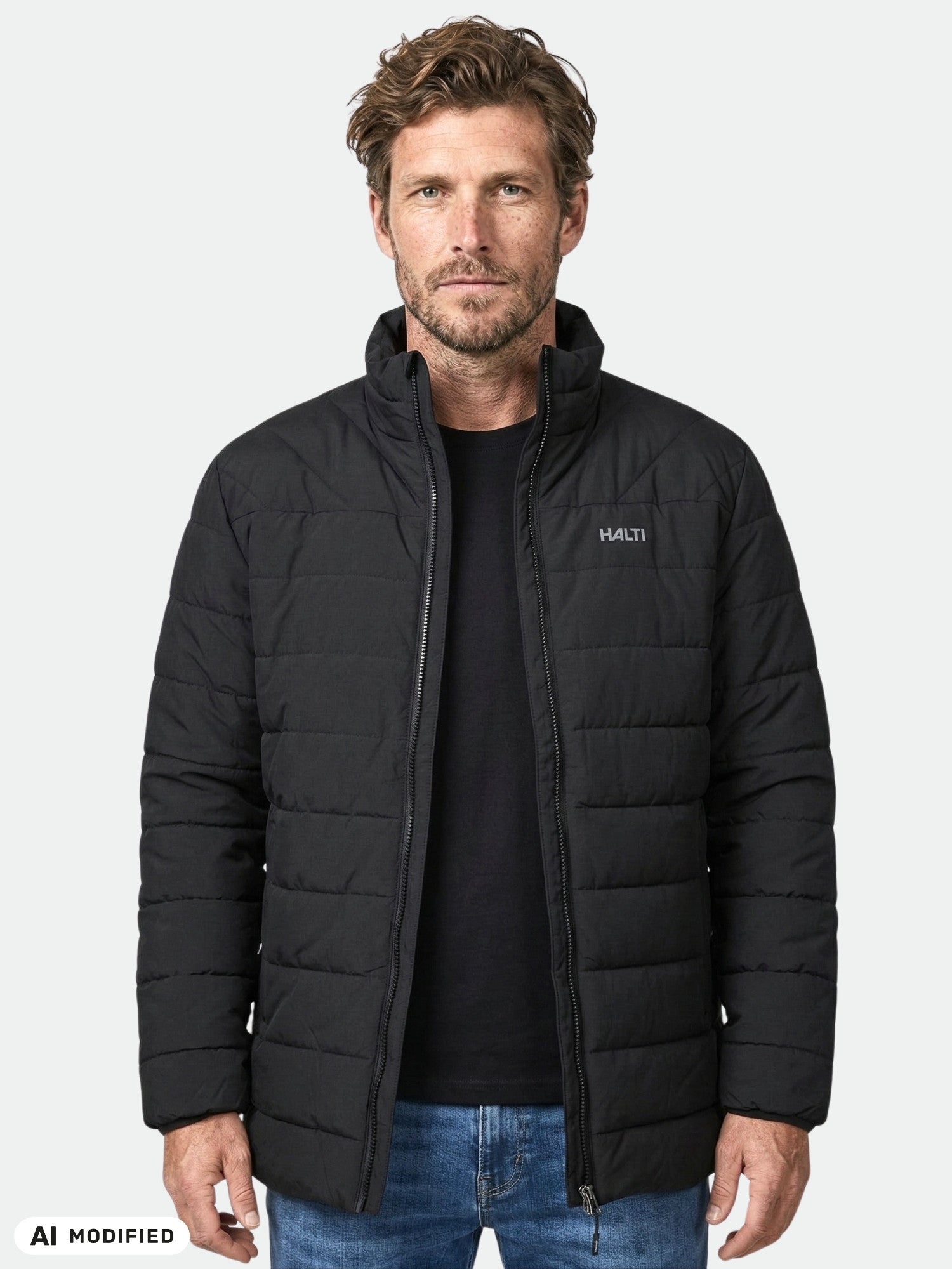 En man står vänd framåt i en svart Halti Haapa Vadderad Jacka Herr, svart skjorta och blå jeans mot en enfärgad bakgrund.