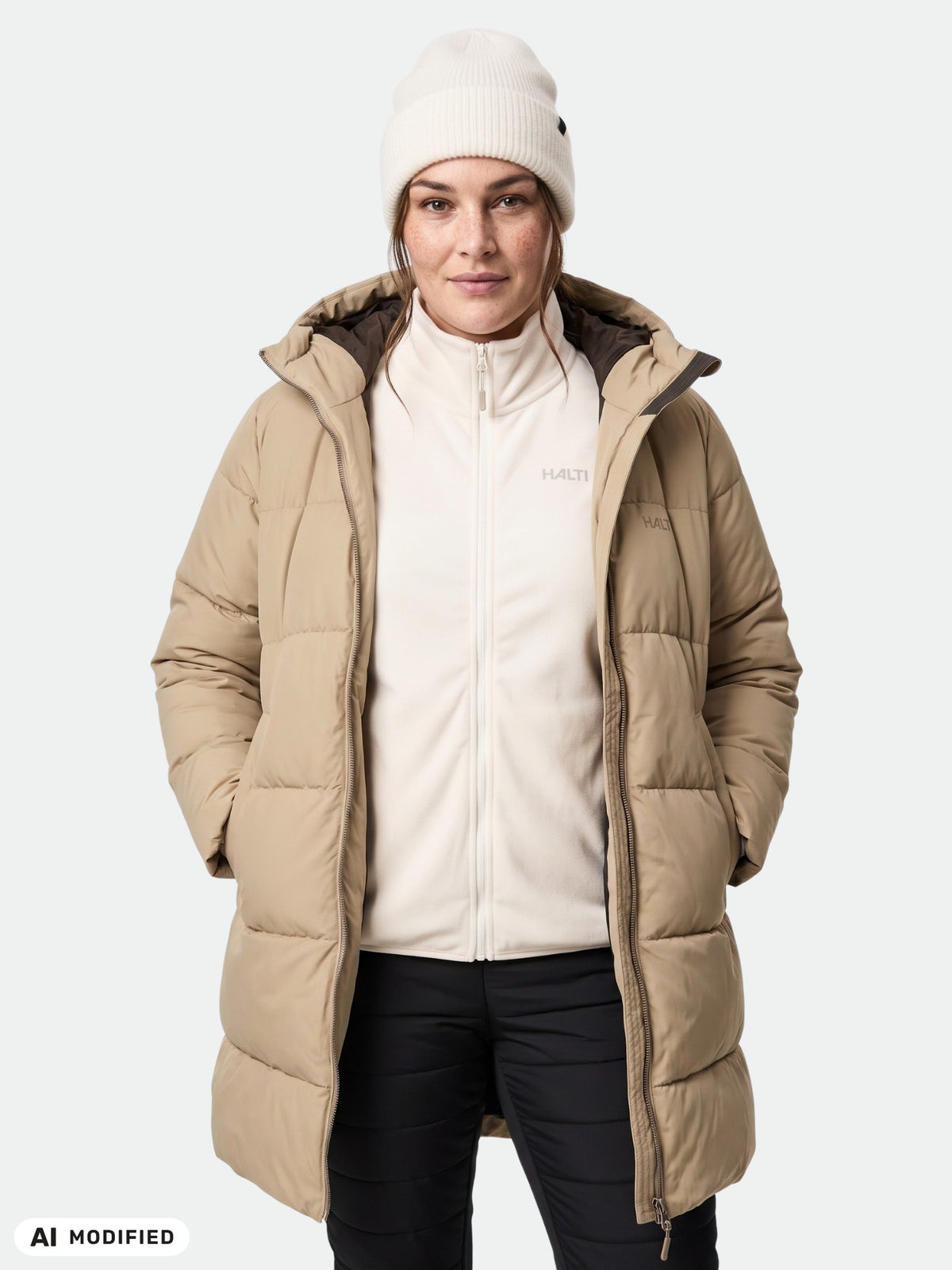 En kvinna bär en Halti Sammal Vadderad Parkajacka Dam Plus i beige med reflekterande detaljer, händerna i fickorna, mot en enfärgad bakgrund.