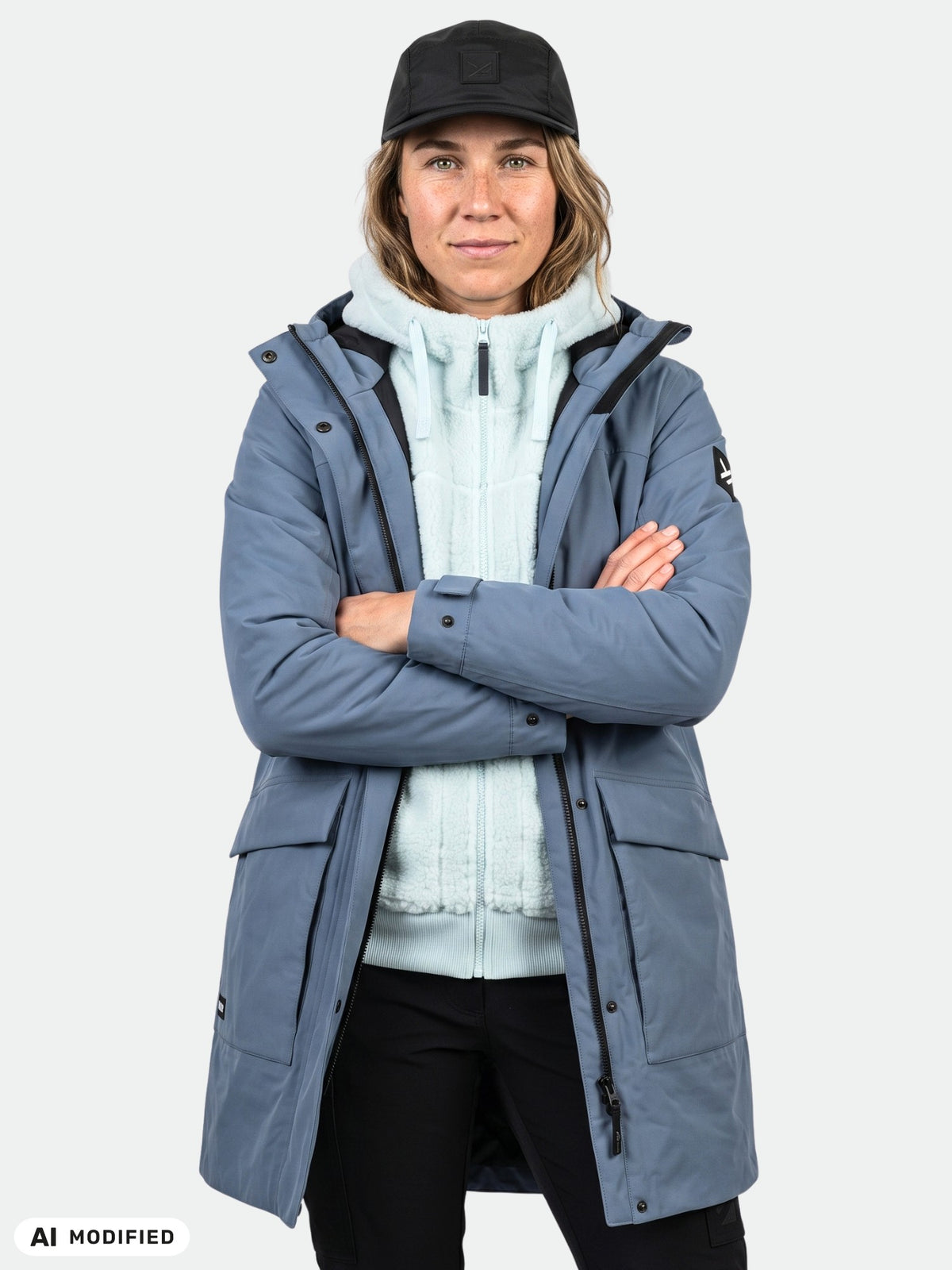 En kvinna klädd i en Halti Kortteli DX-parkas med isolering, ljusblå med dragkedja, svarta byxor och en keps står med armarna i kors.