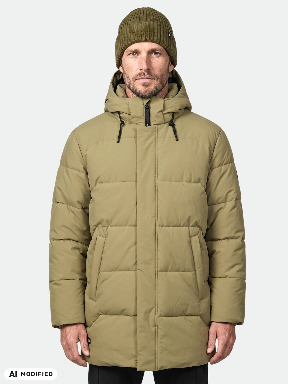 En man i en Halti Brahe Puffer Parka Herr, olivgrön, och en stickad mössa, som står framför en enfärgad ljusgrå bakgrund.
