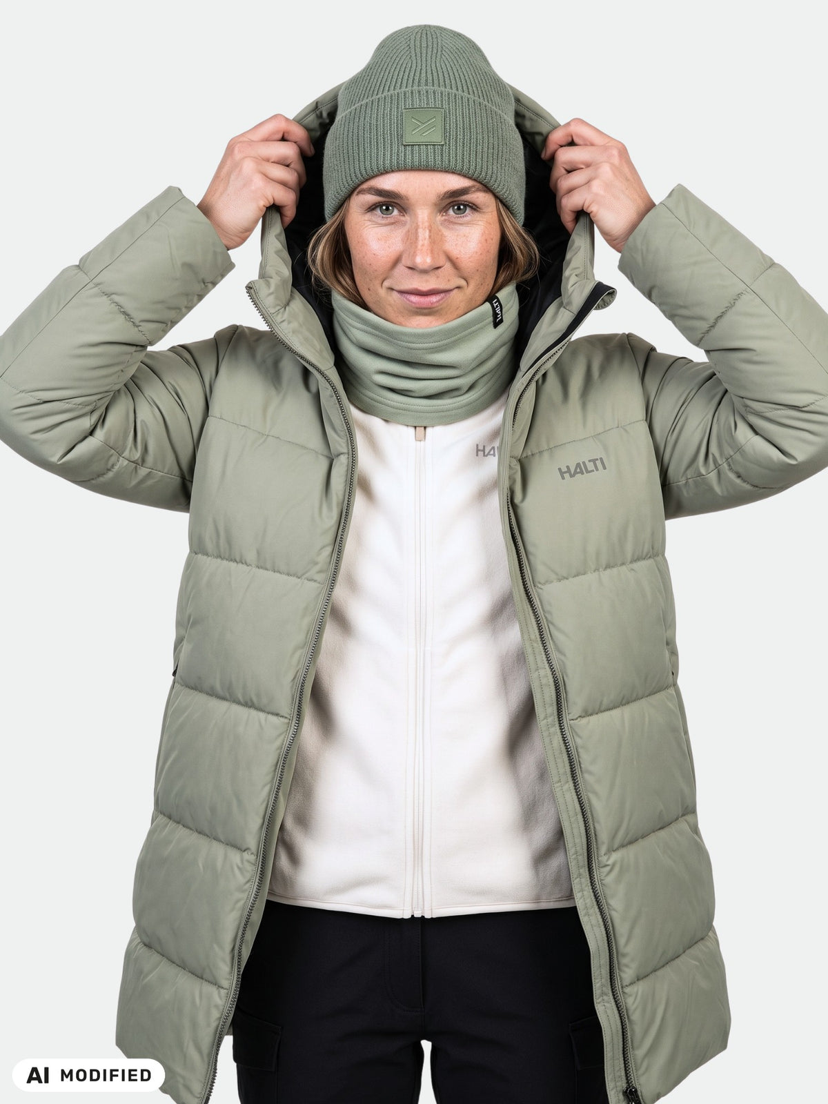 En person i en Halti Leinikko Puffer Parka för dam, salviagrön, med matchande mössa och halsvärmare, som håller i huvan.