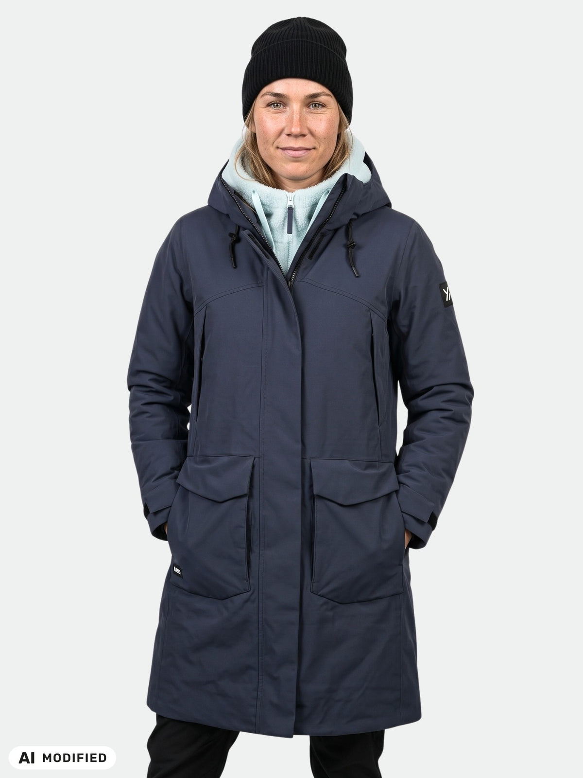 En person klädd i en marinblå Halti Kallio DX Parka Dam, en ljus huvtröja, en svart mössa och svarta byxor framför en enfärgad ljus bakgrund.