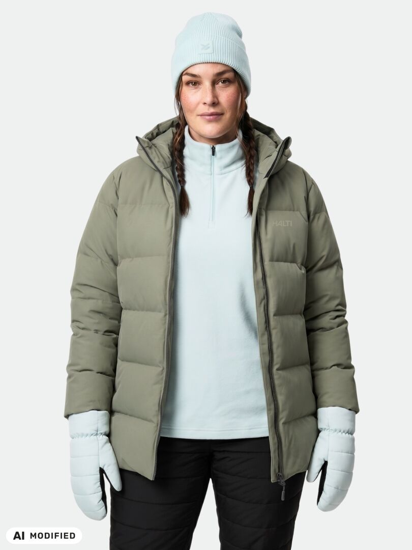 En kvinna klädd i en Leinikko Puffer Parka Dam Plus från Halti, ljusblå handskar och en mössa står framför en enfärgad bakgrund.