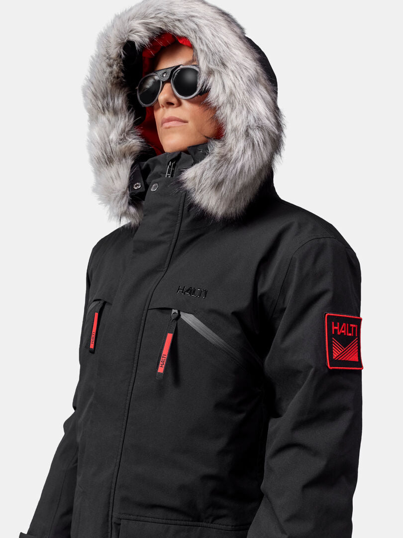 Person klädd i Halti 1976 Down Parka Unisex från Halti med pälsfodrad huva, solglasögon och röda detaljer på ärmarna.