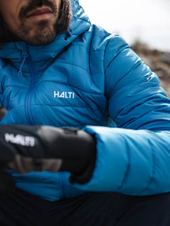 En man i blå Halti Huippu Dunjacka Herr med handskar sitter utomhus, Pertex Quantum-tyg syns, bakgrunden är suddig.