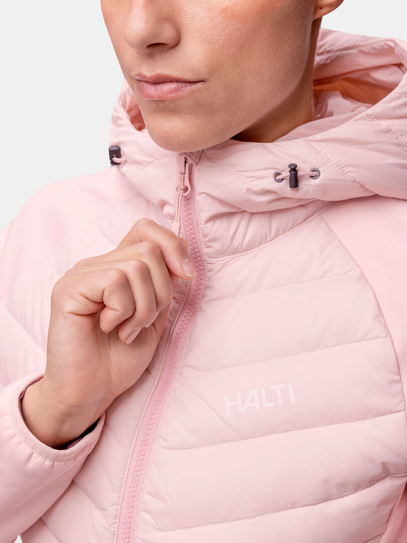 Person som drar upp dragkedjan på Halti Huippu Hybrid Dunjacka Dam i ljusrosa; logotyp och Pertex Quantum ripstop-tyg syns.